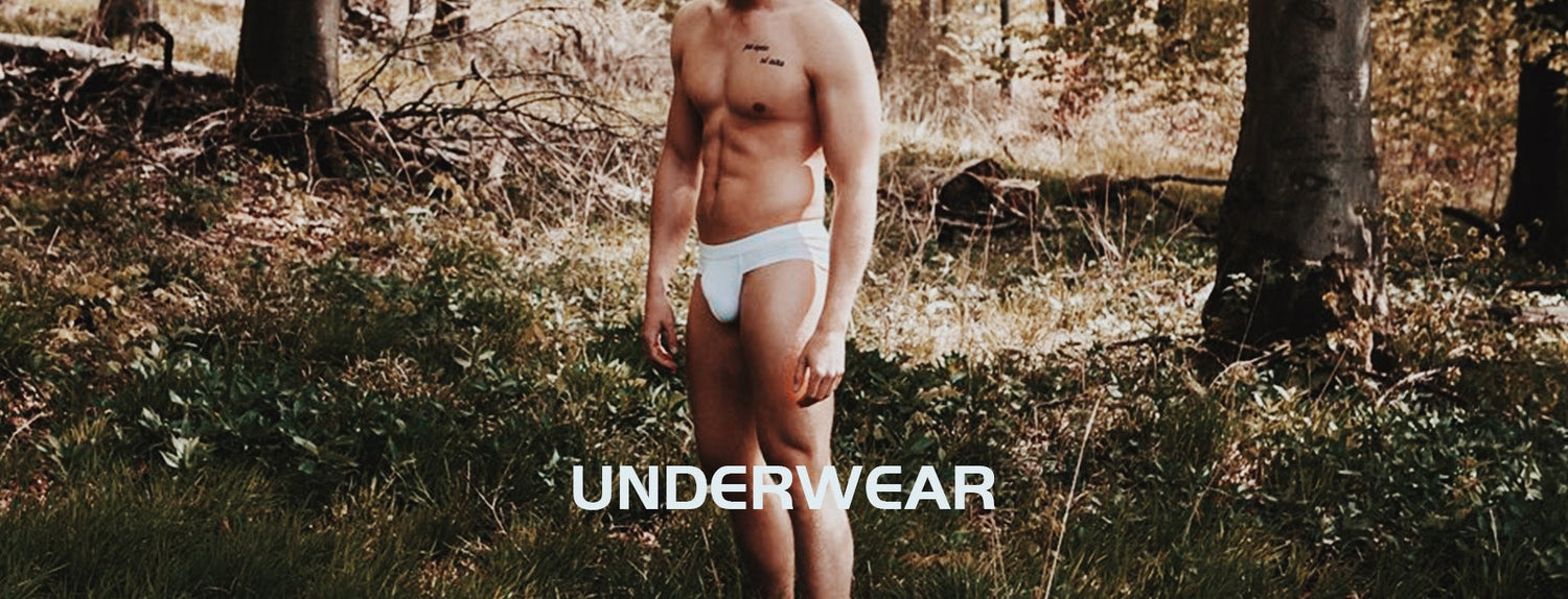 A male model wearing Furqx white men's briefs stands in an outdoor forest environment.